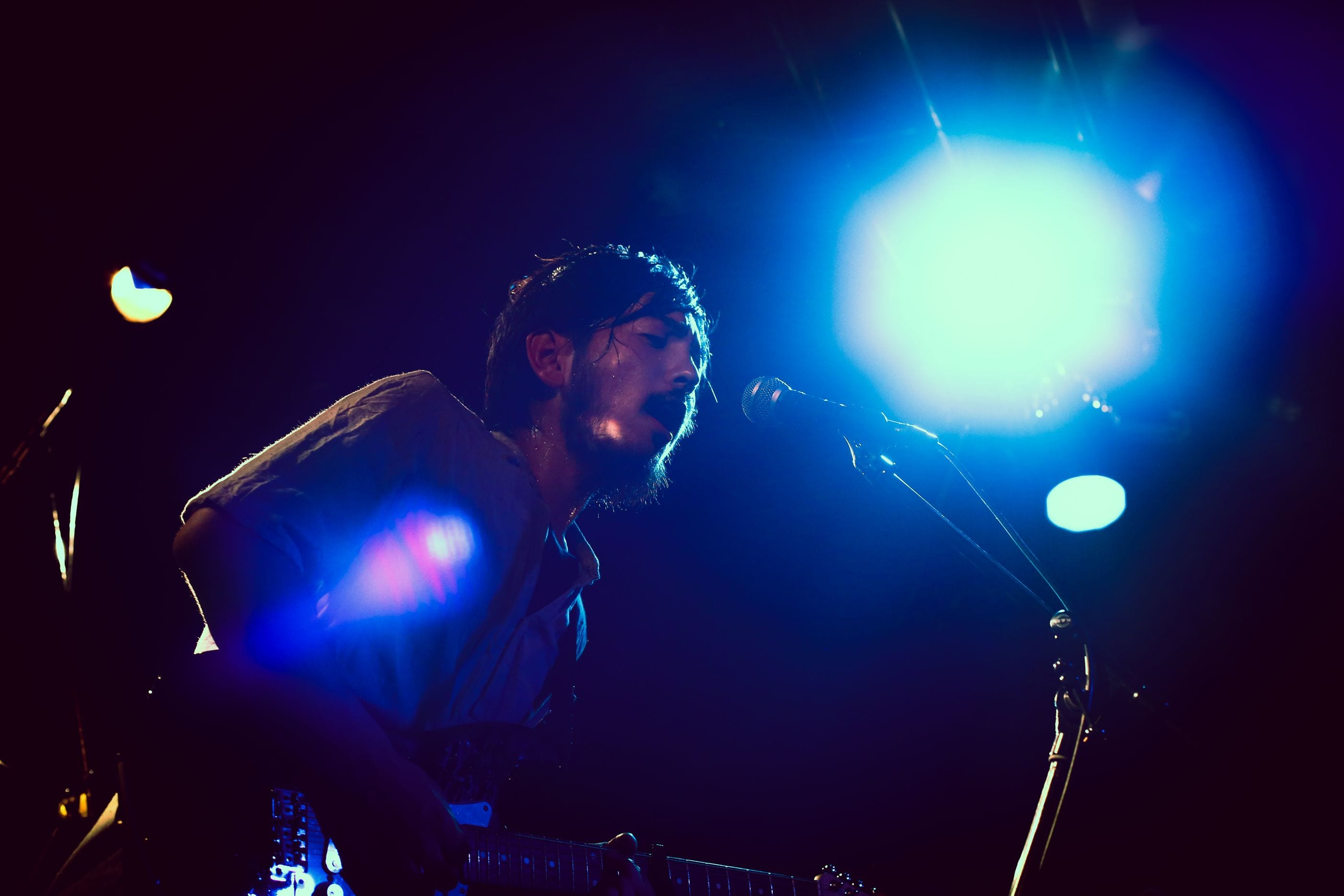 Potomelli on a dark stage under a blue spotlight, singing into the microphone while playing guitar.