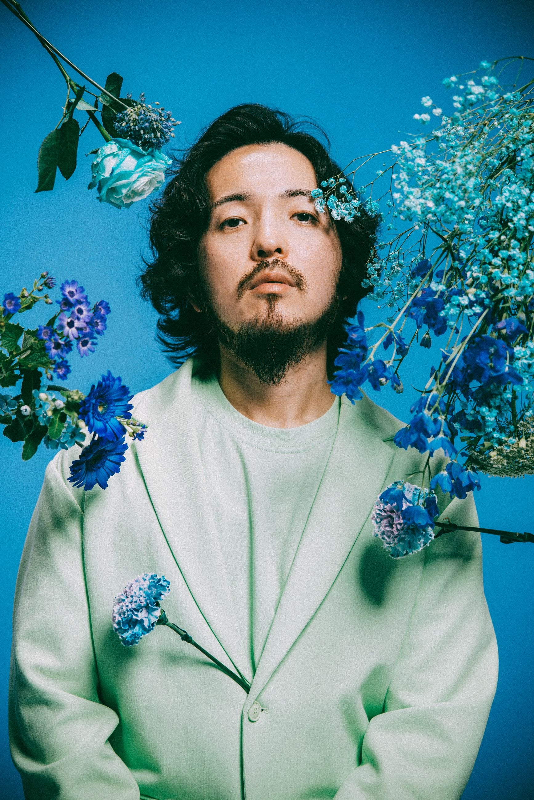 Potomelli in a soft green suit against electric blue, surrounded by blue delphiniums, roses and baby’s breath.